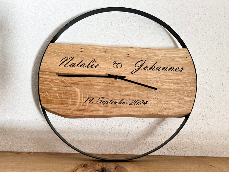 Wanduhr aus massivem Eichenholz mit schwarzem Metallring. Im Holz sind die Namen des Brautpaares und das Hochzeitsdatum graviert. Es dient als Hochzeitsgeschenk.