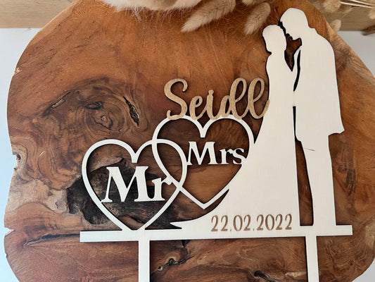 Auf dem Bild ist ein cake topper zur Hochzeit aus Holz abgebildet. Mit einem Brautpaar, Mr und Ms der Nachname des Paares und das Hochzeitsdatum graviert.