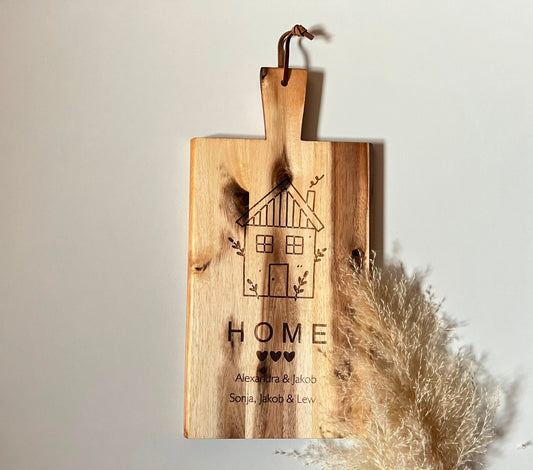 Graviertes Holz-Schneidebrett mit einem Hausmotiv und den Namen der Familienmitglieder. Persönliches Geschenk zum Einzug oder als Hochzeitsgeschenk.